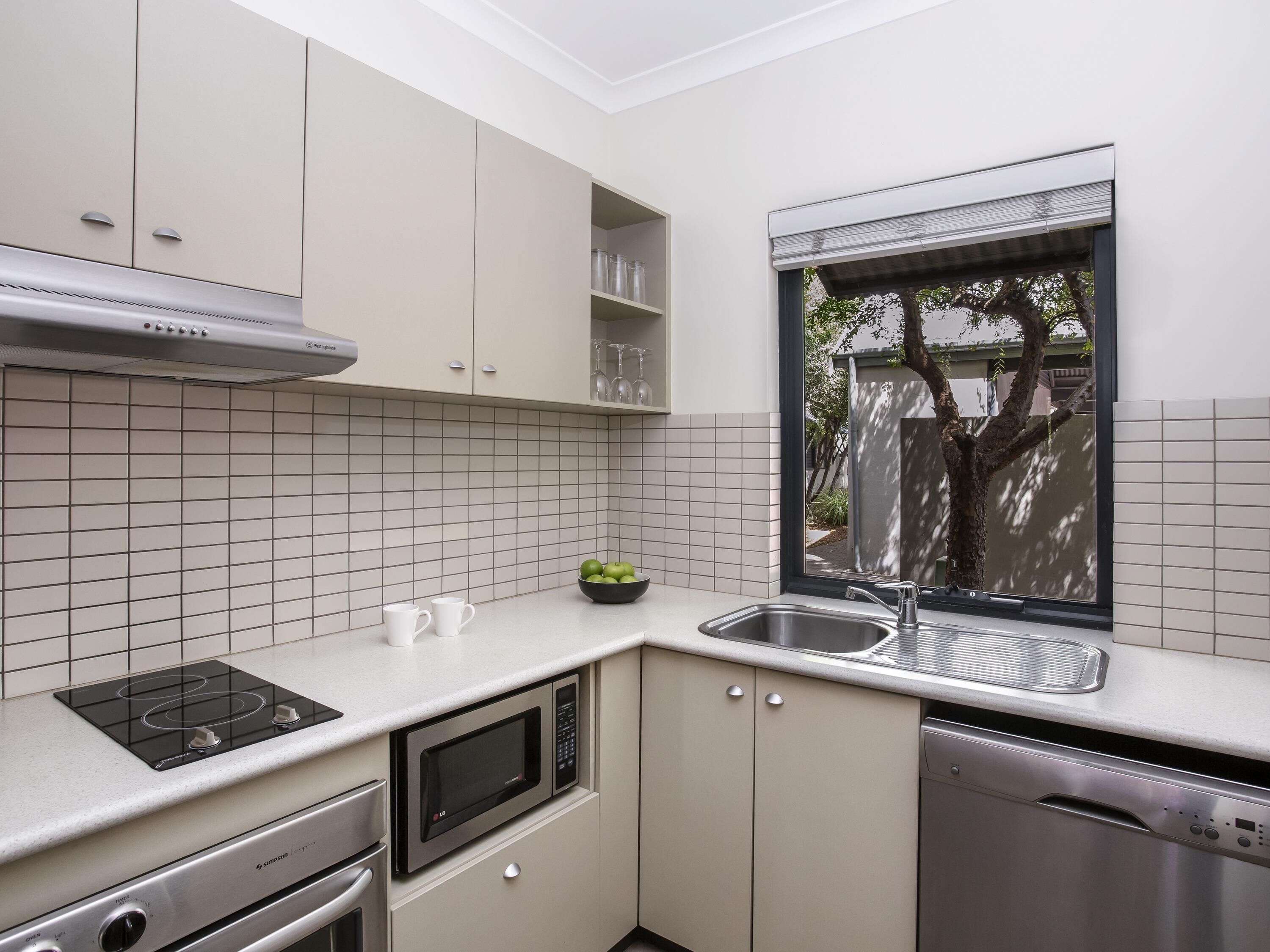 A compact kitchen with a built-in microwave, stove, and dishwasher, a window with a view of greenery, and a tiled backsplash.