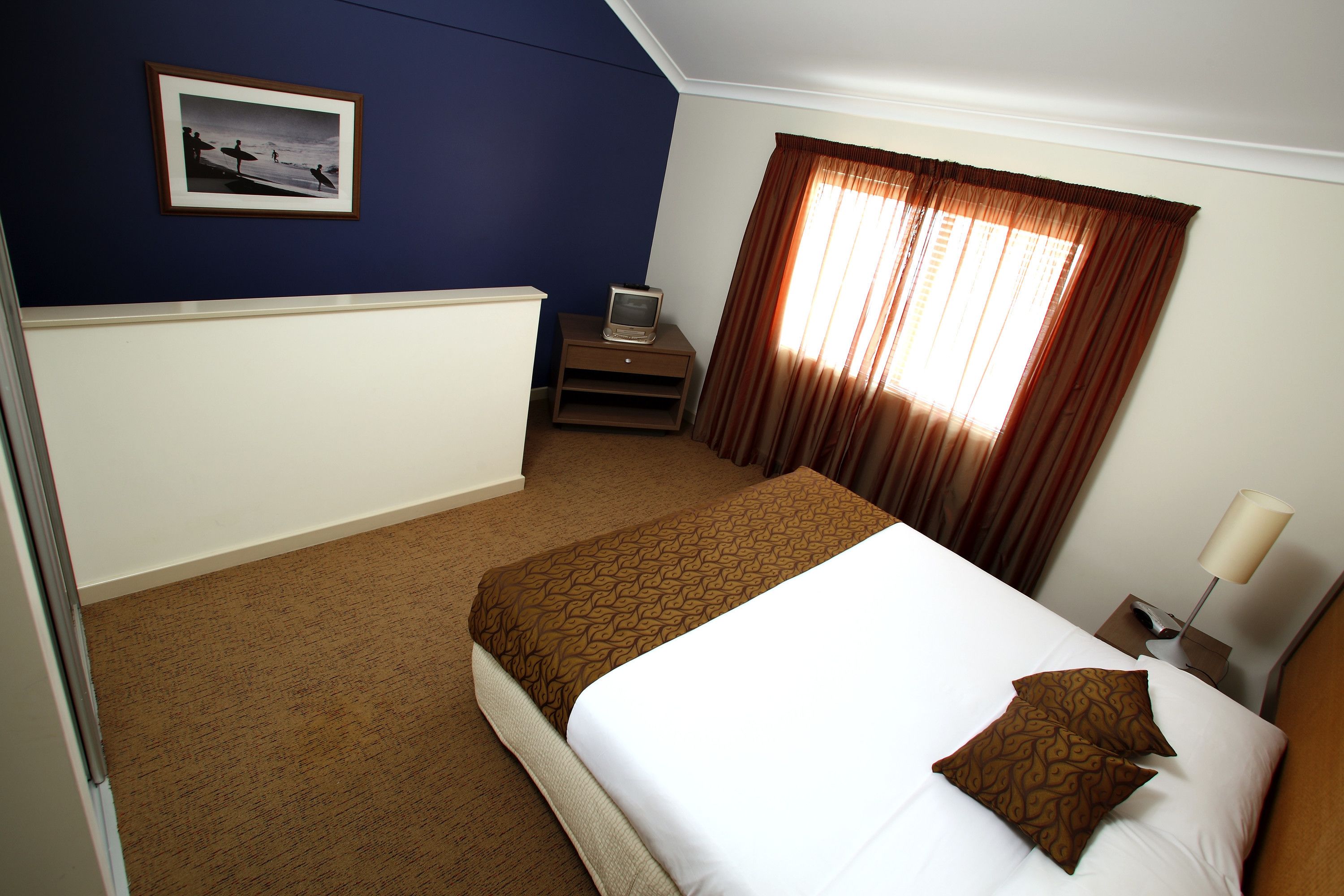 A hotel room with a bed, a TV on a stand, a window with curtains, and a framed picture on the wall.