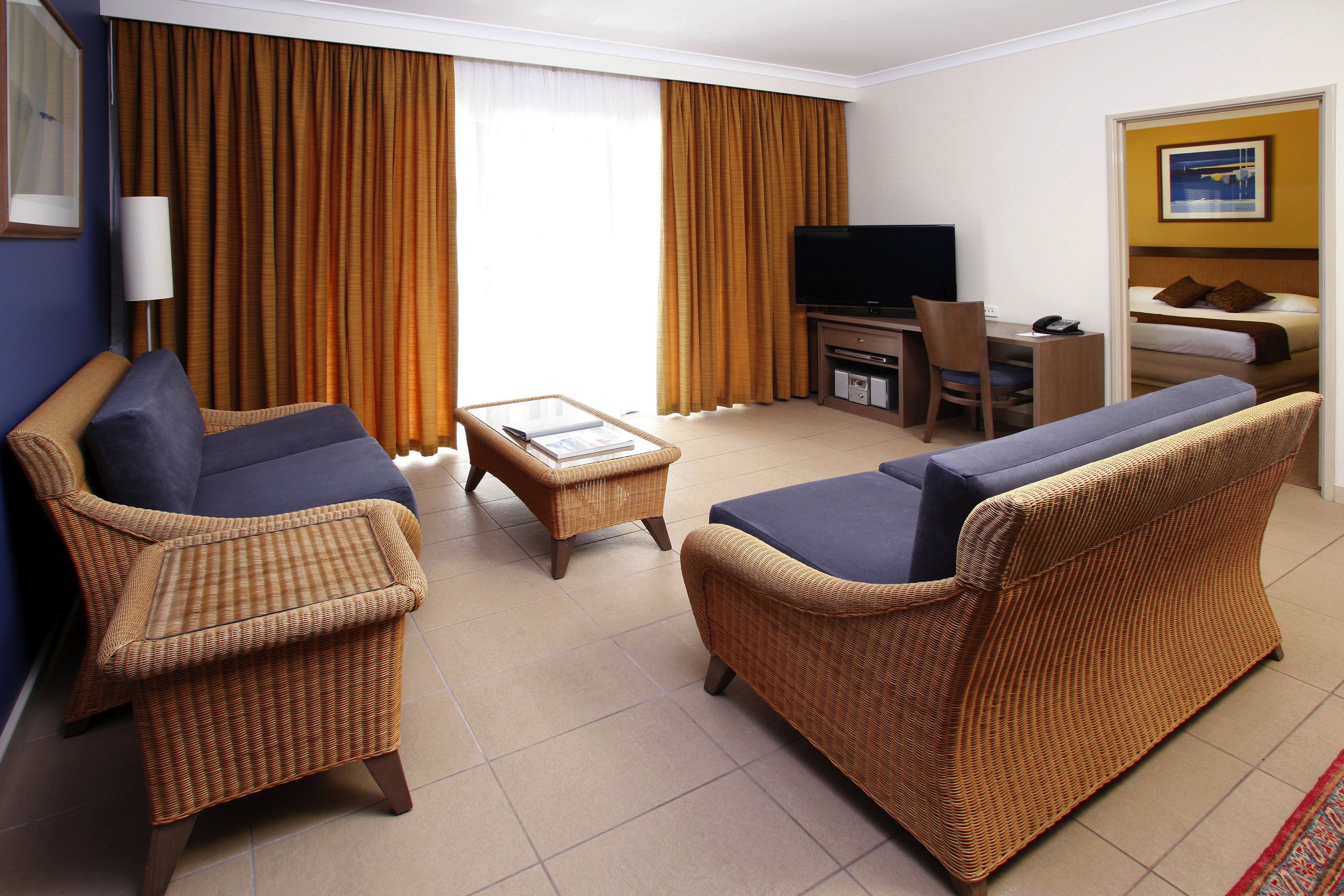 Quarto de hotel com sofá, poltrona e uma pequena mesa. Há uma televisão e uma escrivaninha com cadeira.