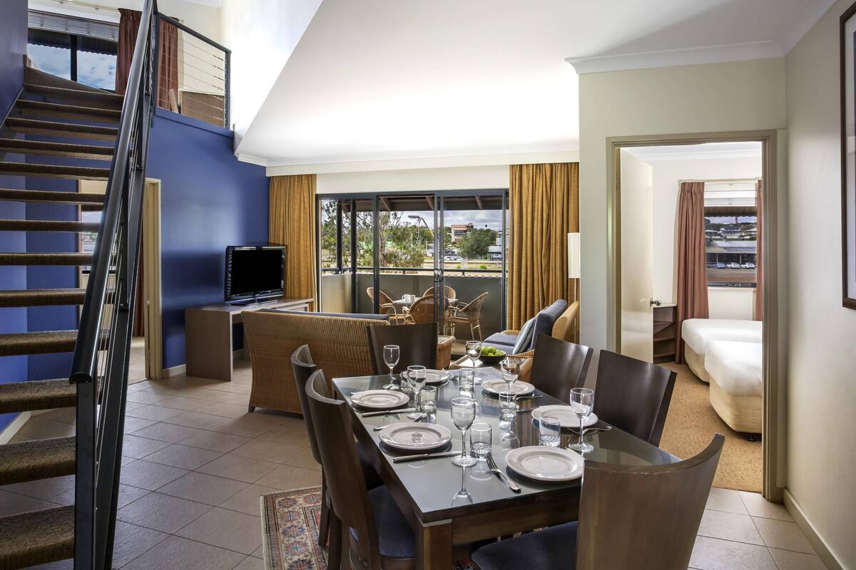 Uma área de jantar com mesa posta para quatro pessoas, televisão e varanda com mobiliário de exterior.
