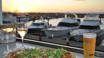 A pizza with a glass of white wine and a beer on a table with a marina view in the background.