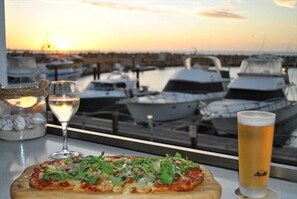 Pizza con una copa de vino blanco y una cerveza sobre una mesa, con vista al puerto deportivo al fondo.
