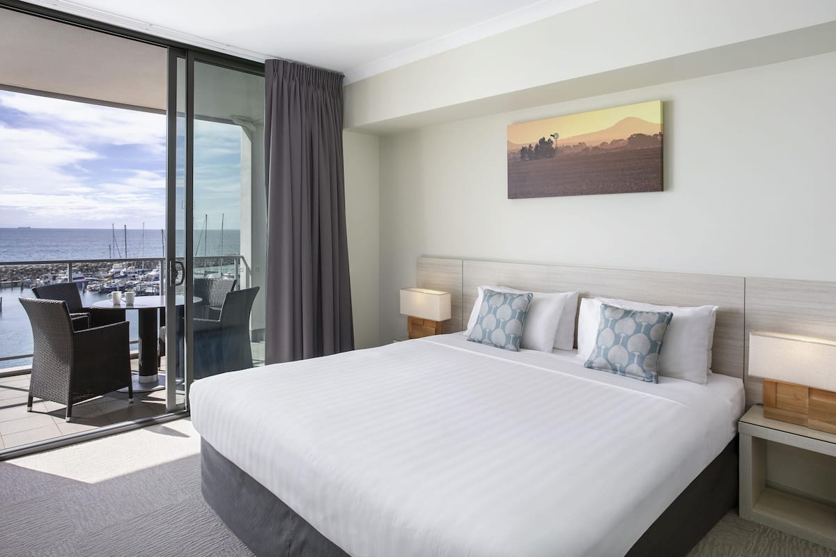 Quarto de hotel com uma cama grande, duas mesas de cabeceira, vista para o porto de iates e um quadro na parede.