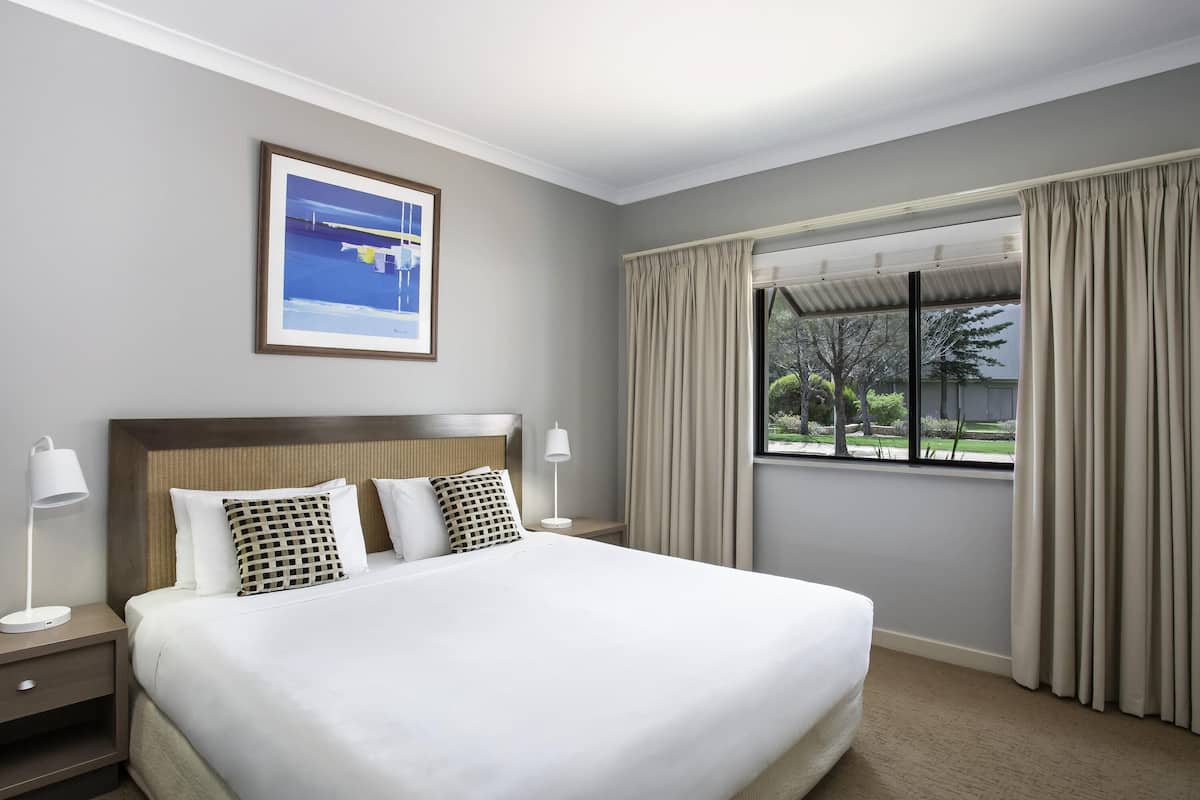 Quarto de hotel com uma cama grande, uma mesa de cabeceira, uma luminária, um quadro na parede e uma janela com cortinas.