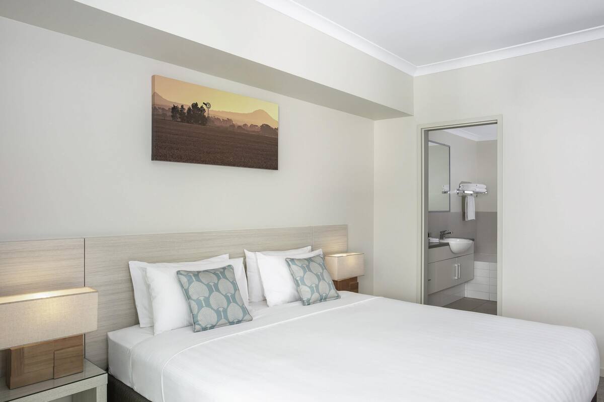 Quarto de hotel com cama, abajures, um quadro, banheiro com espelho e pia.
