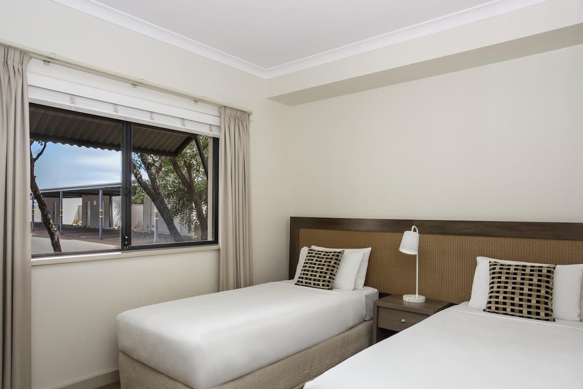Quarto de hotel com duas camas, uma mesinha de cabeceira, uma luminária e uma janela com cortinas.