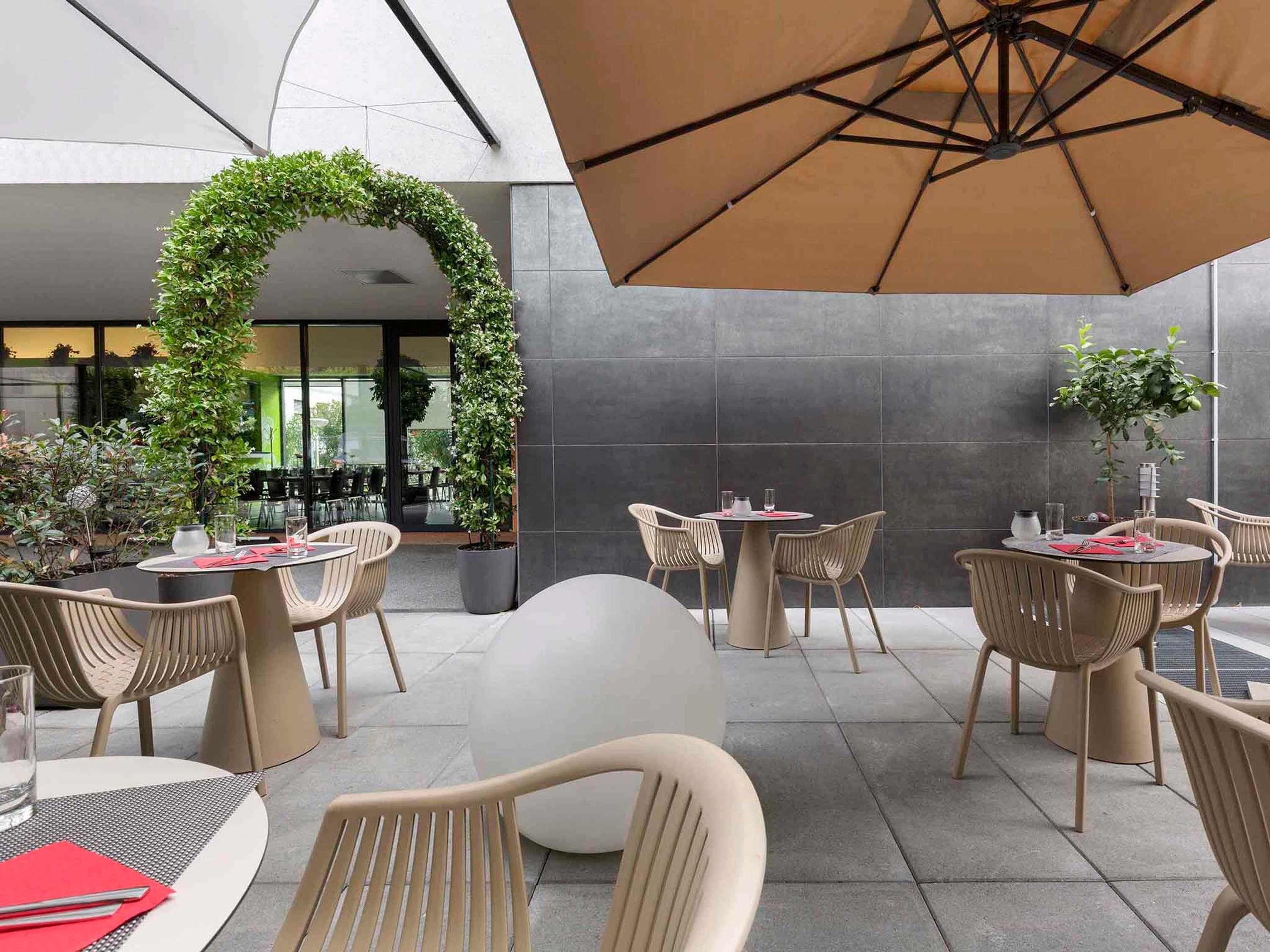 An outdoor seating area with round tables and chairs, a large umbrella, and a green archway.