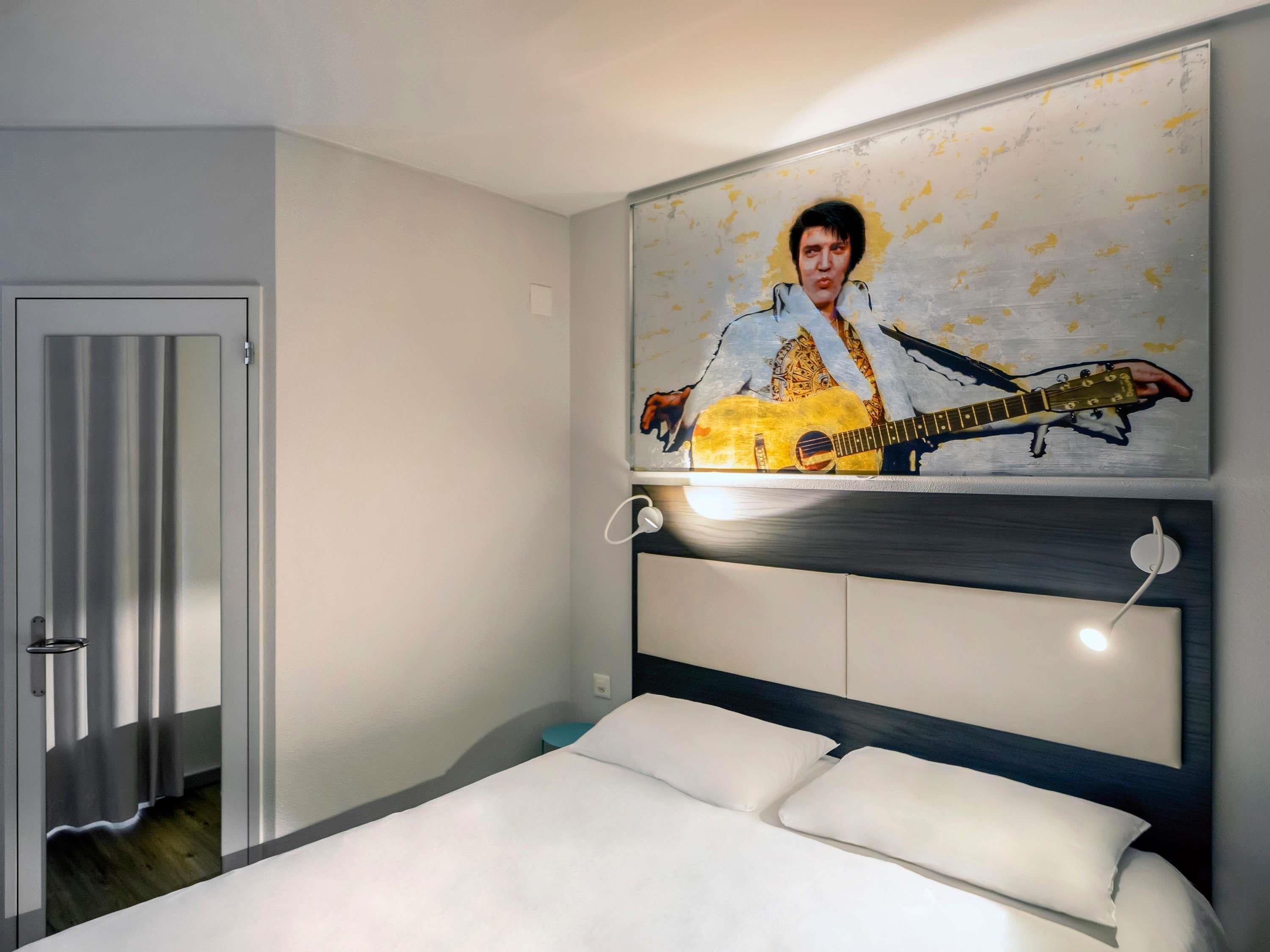 Ein Schlafzimmer mit Bett, einem Kopfteil, einem an der Wand hängenden Bild einer Gitarre und einer Tür.