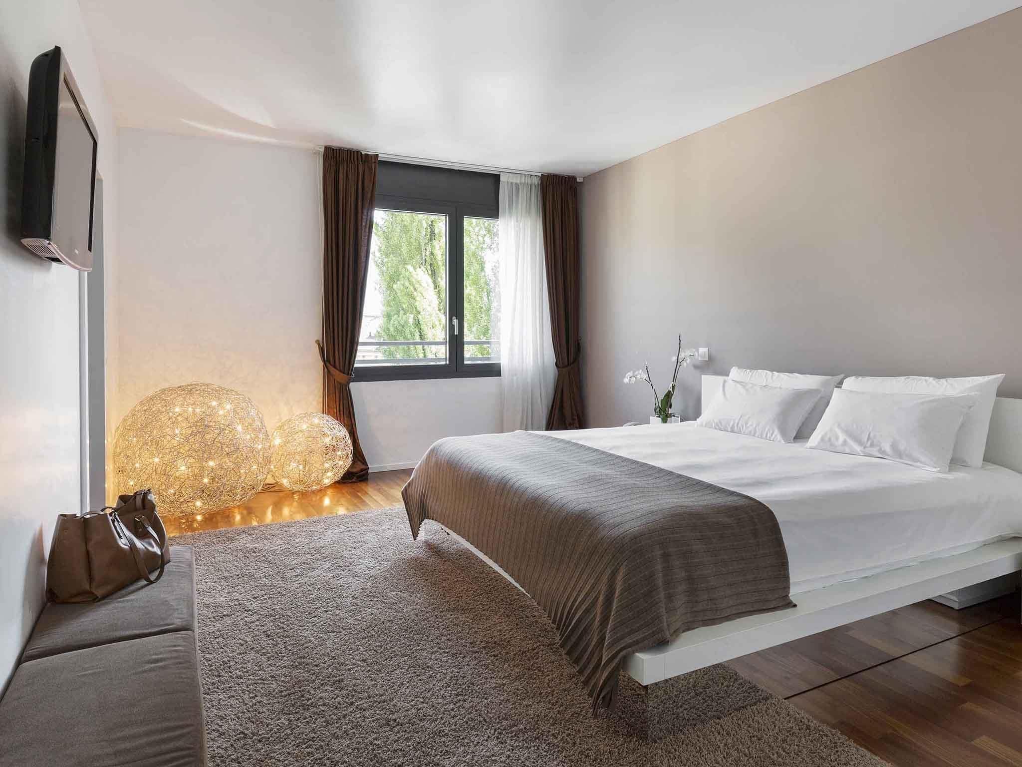 A modern bedroom with a large bed, a flat-screen TV mounted on the wall, a window with curtains, and a decorative light fixture.