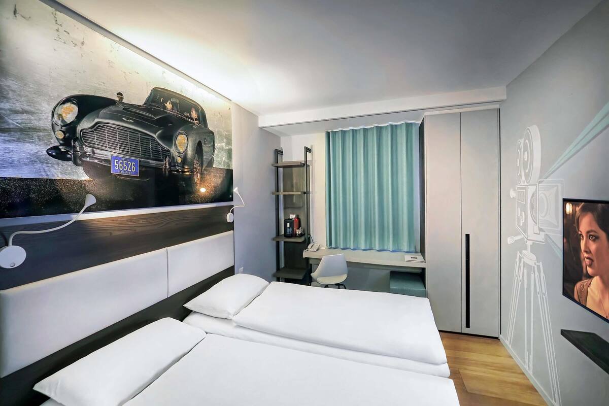 Una habitación de hotel moderna con una cama grande, un escritorio con silla, un armario y un televisor montado en la pared.