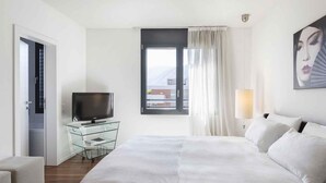 Una habitación de hotel con cama, televisor en un mueble de vidrio, ventana con cortinas y un cuadro en la pared.