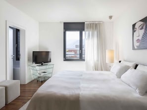 Una habitación de hotel con cama, televisor en un mueble de vidrio, ventana con cortinas y un cuadro en la pared.