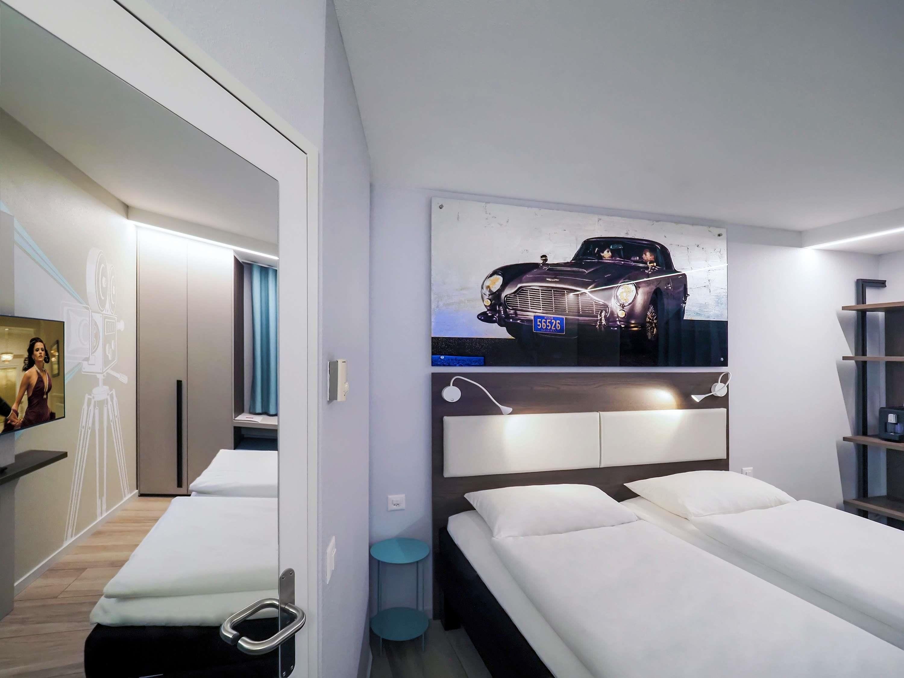A modern hotel room with a large bed, a headboard with integrated lighting, a wall-mounted TV, and a framed picture of a classic car.