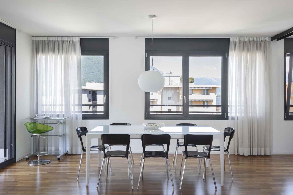 Un comedor moderno con una mesa blanca y sillas negras, ventanas amplias con cortinas, y vistas a edificios y montañas.