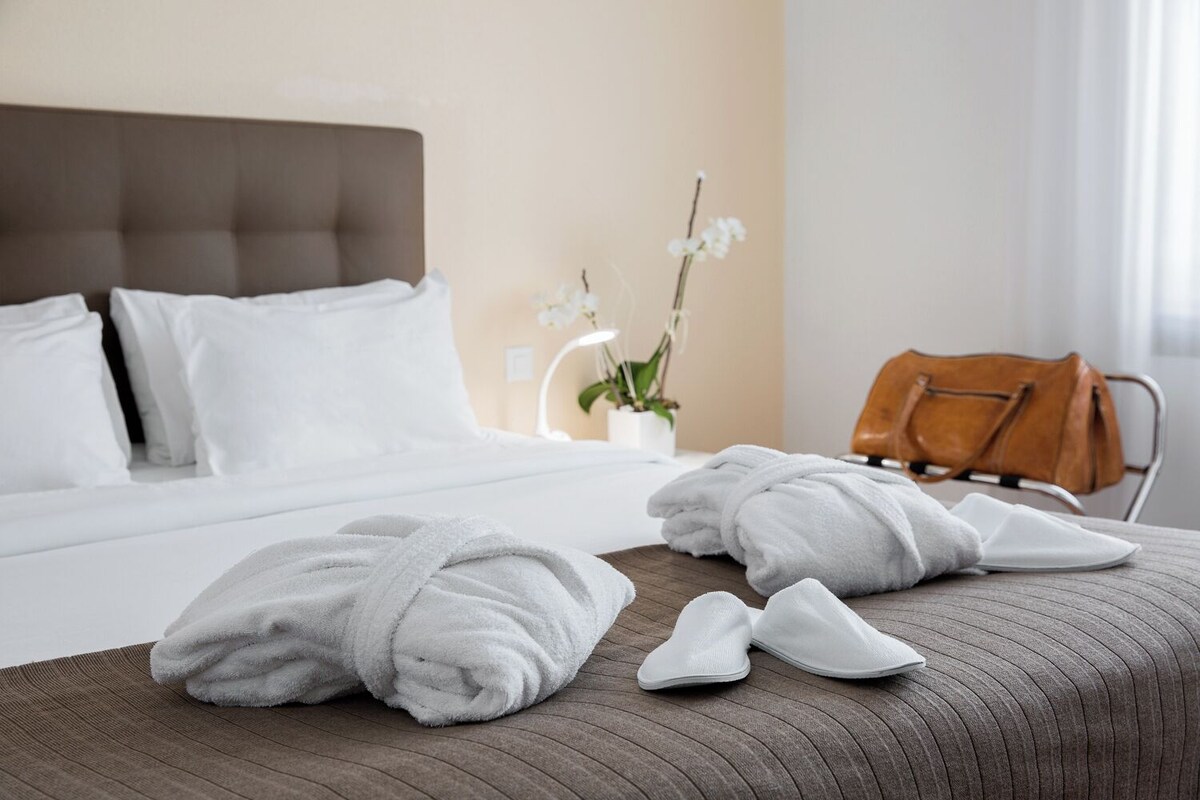 Una habitació d'hotel amb un llit ben fet, una taula de nit de fusta amb tovalloles i xalets, una cadira de cuir marró i una orquídia en test.