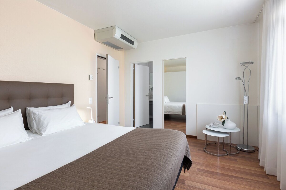 Una habitació d'hotel amb un llit gran, una taula de nit, un mirall, una dutxa i un aparell d'aire condicionat.
