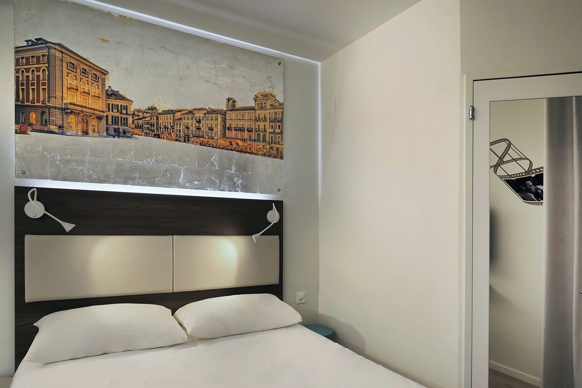 Habitació d'hotel amb un llit, un panell de capçalera amb un mural de paisatge urbà, una taula de nit i una porta decorada amb una franja de pel·lícula.