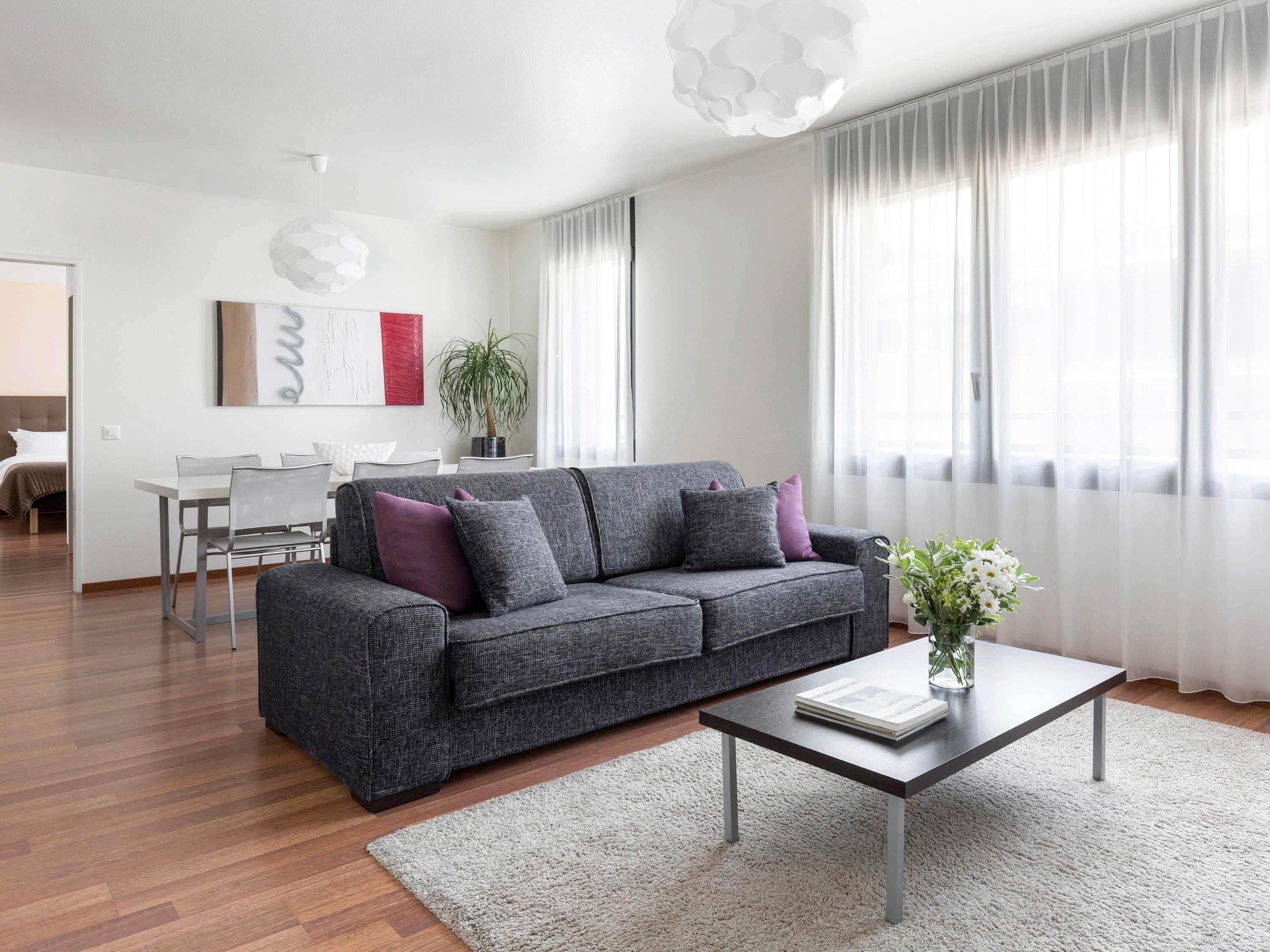 Un salon moderne avec un canapé gris, une table basse et, au fond, un coin repas.