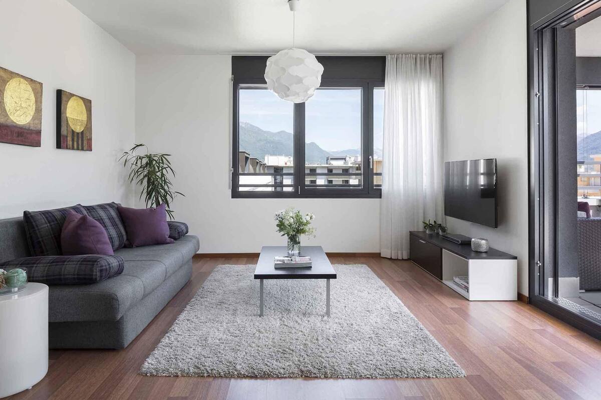 Una sala de estar moderna con un sofá gris, una mesa de centro, un televisor y un ventanal con vistas a edificios y montañas.