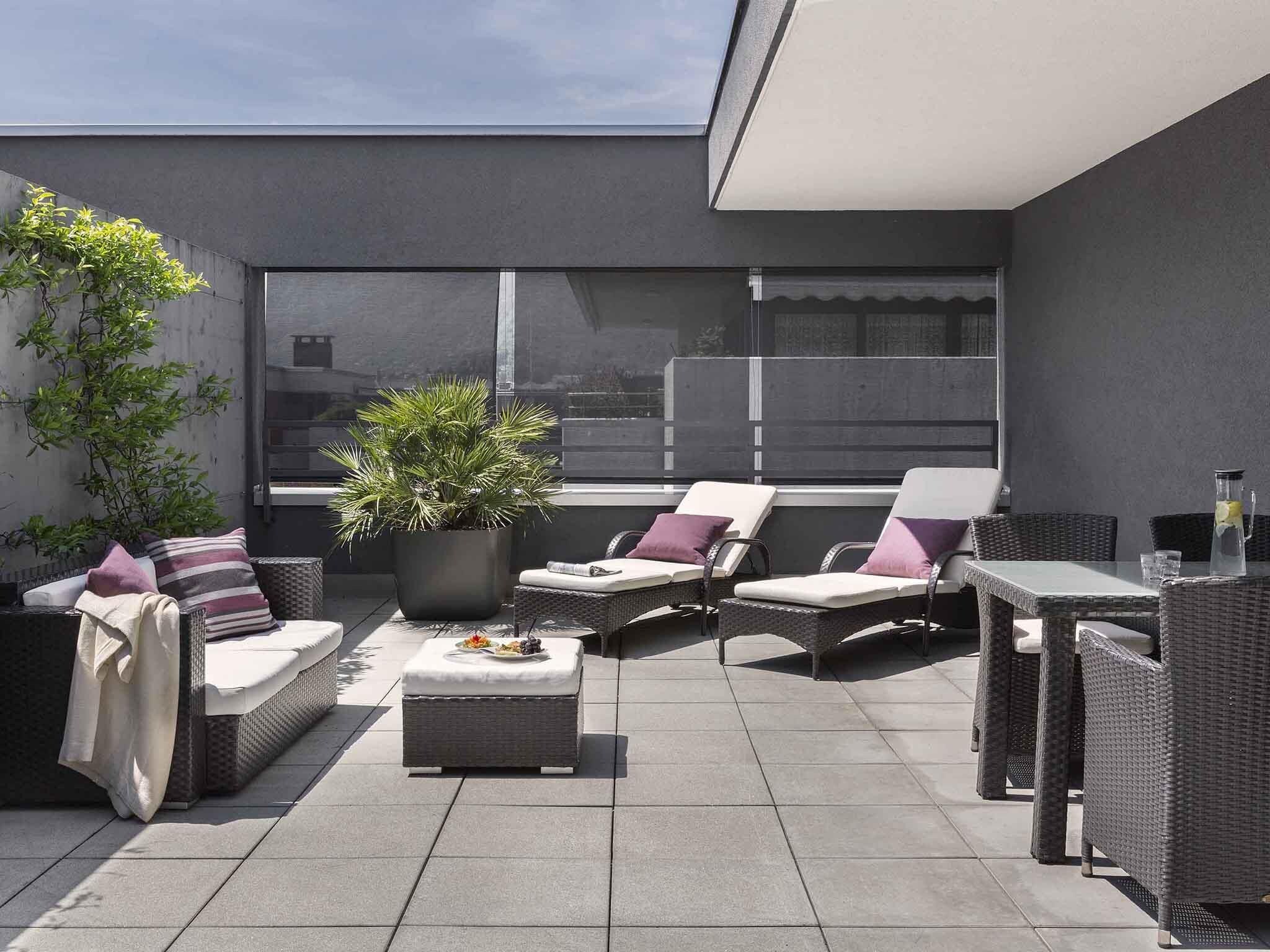 A rooftop terrace with wicker furniture, a potted plant, and a table with a drink.