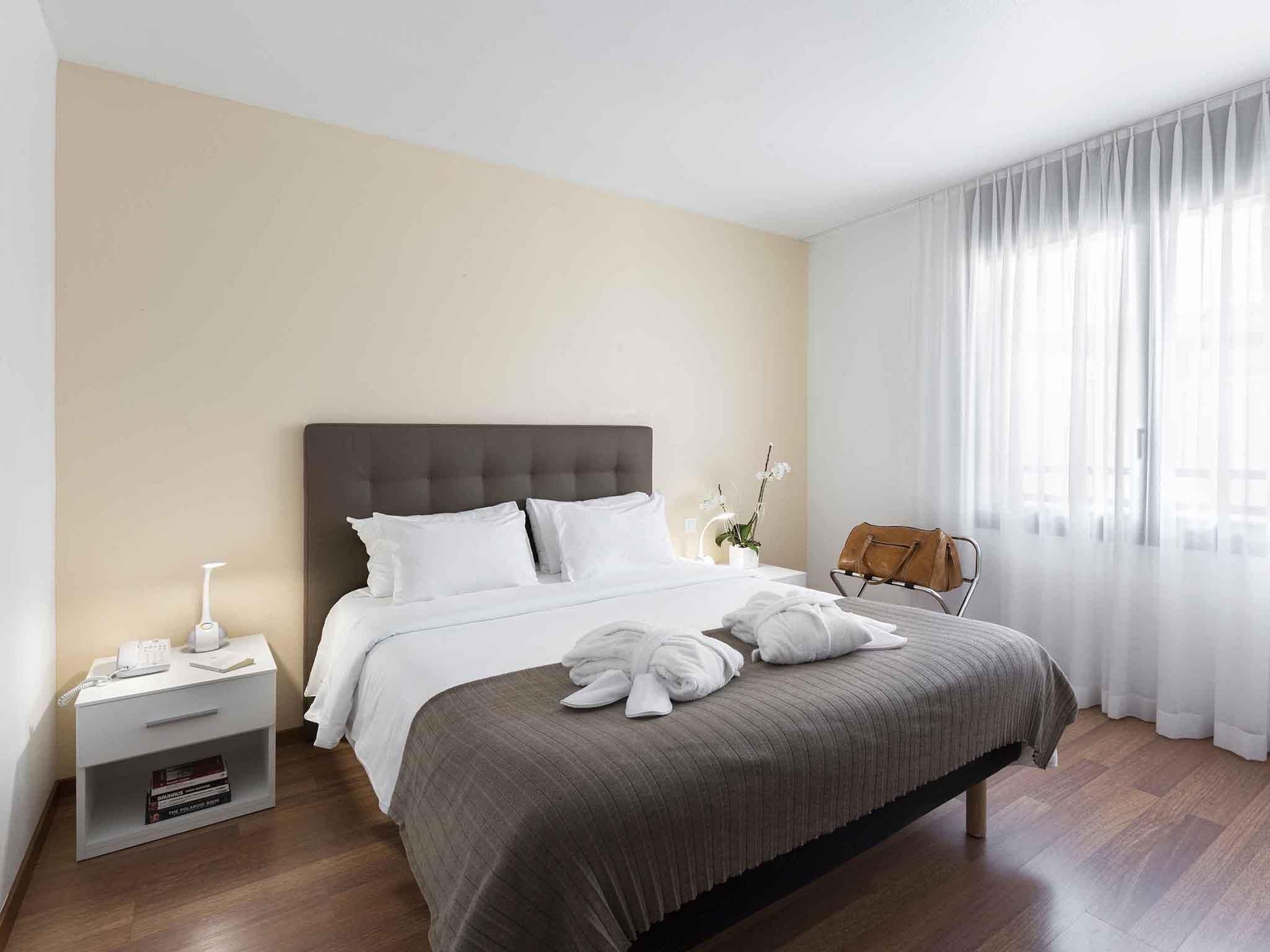 Ein Hotelzimmer mit Bett, Nachttisch, Lampe und Blick durch durchsichtige Vorhänge nach draußen.