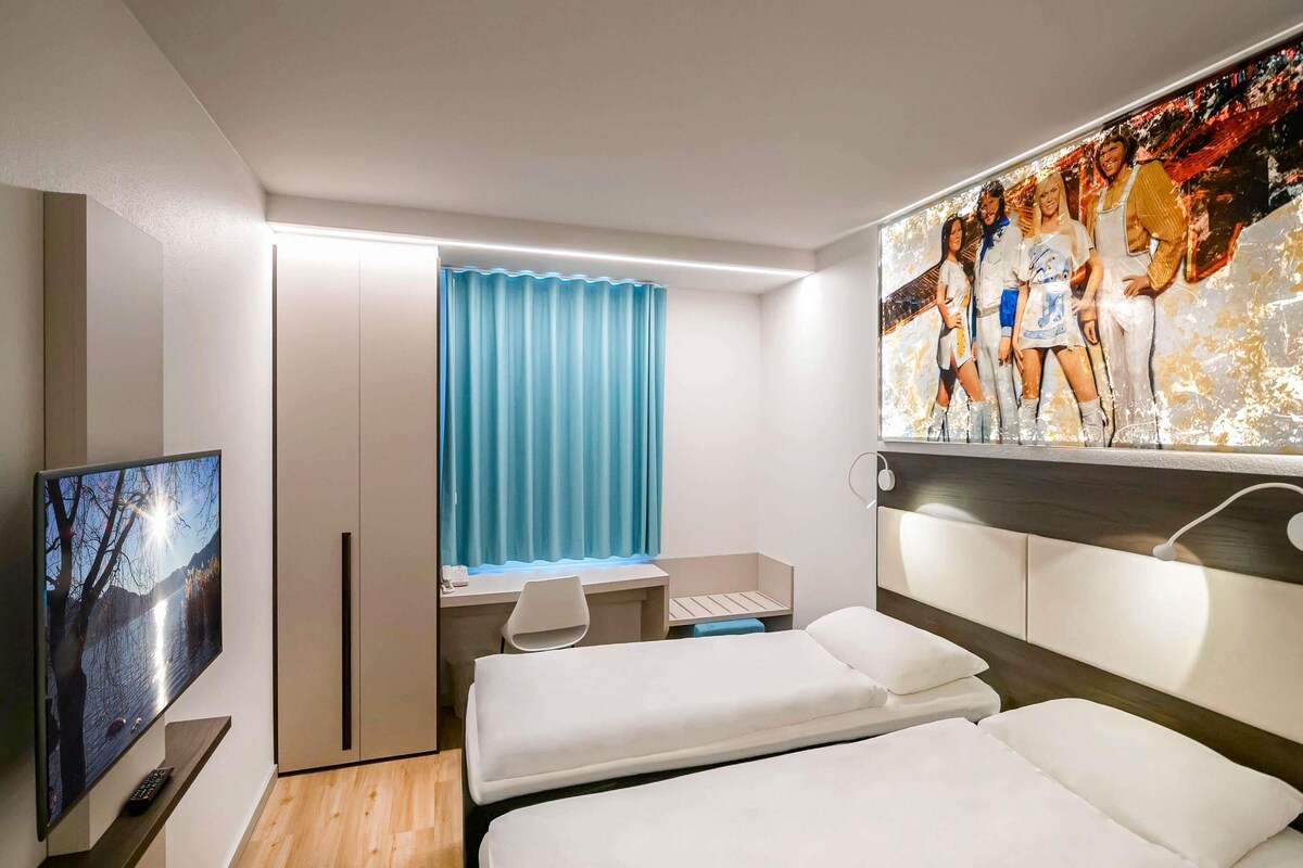 Habitación de hotel con dos camas, un escritorio, una silla, un televisor y un cuadro grande en la pared.