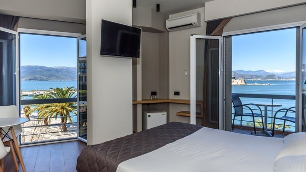 Ein Hotelzimmer mit Bett, Fernseher, Schreibtisch und einem Balkon mit Blick auf Meer und Berge.