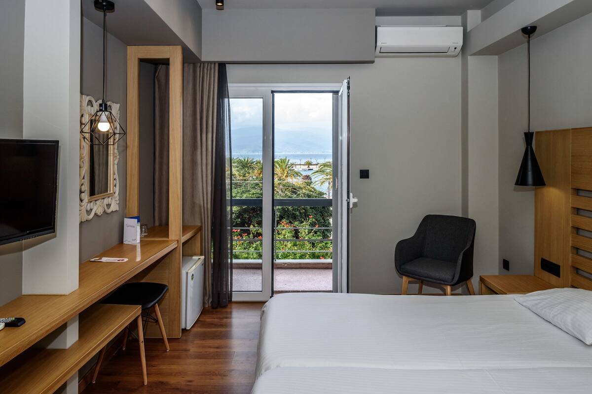 A hotel room with a bed, a desk, a chair, a TV, and a balcony with a view of the sea.