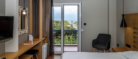 A hotel room with a bed, a desk with a chair, a TV, a balcony with a view of the sea, and an air conditioning unit.