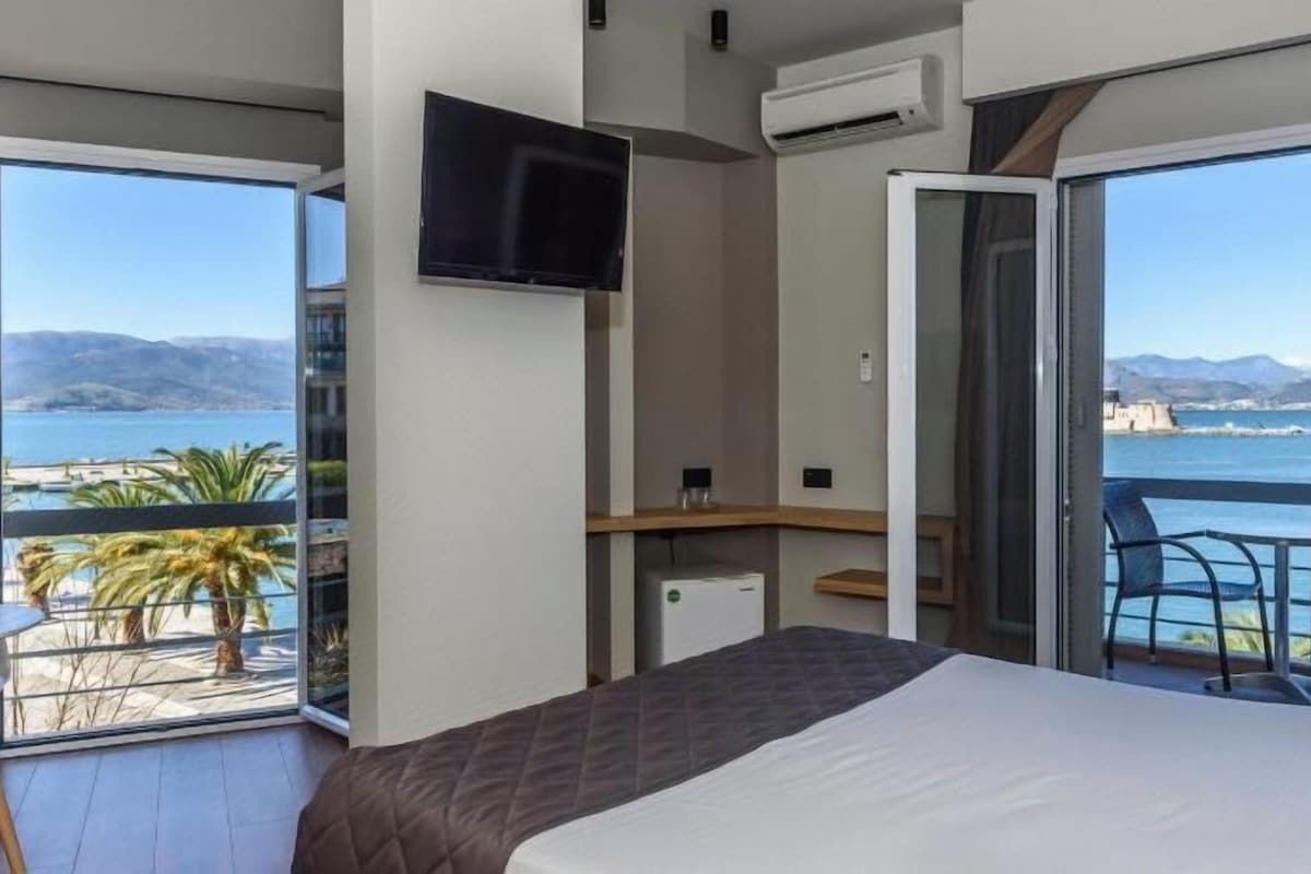 A bedroom with a bed, a desk, and a TV. There is a balcony with a view of the sea.