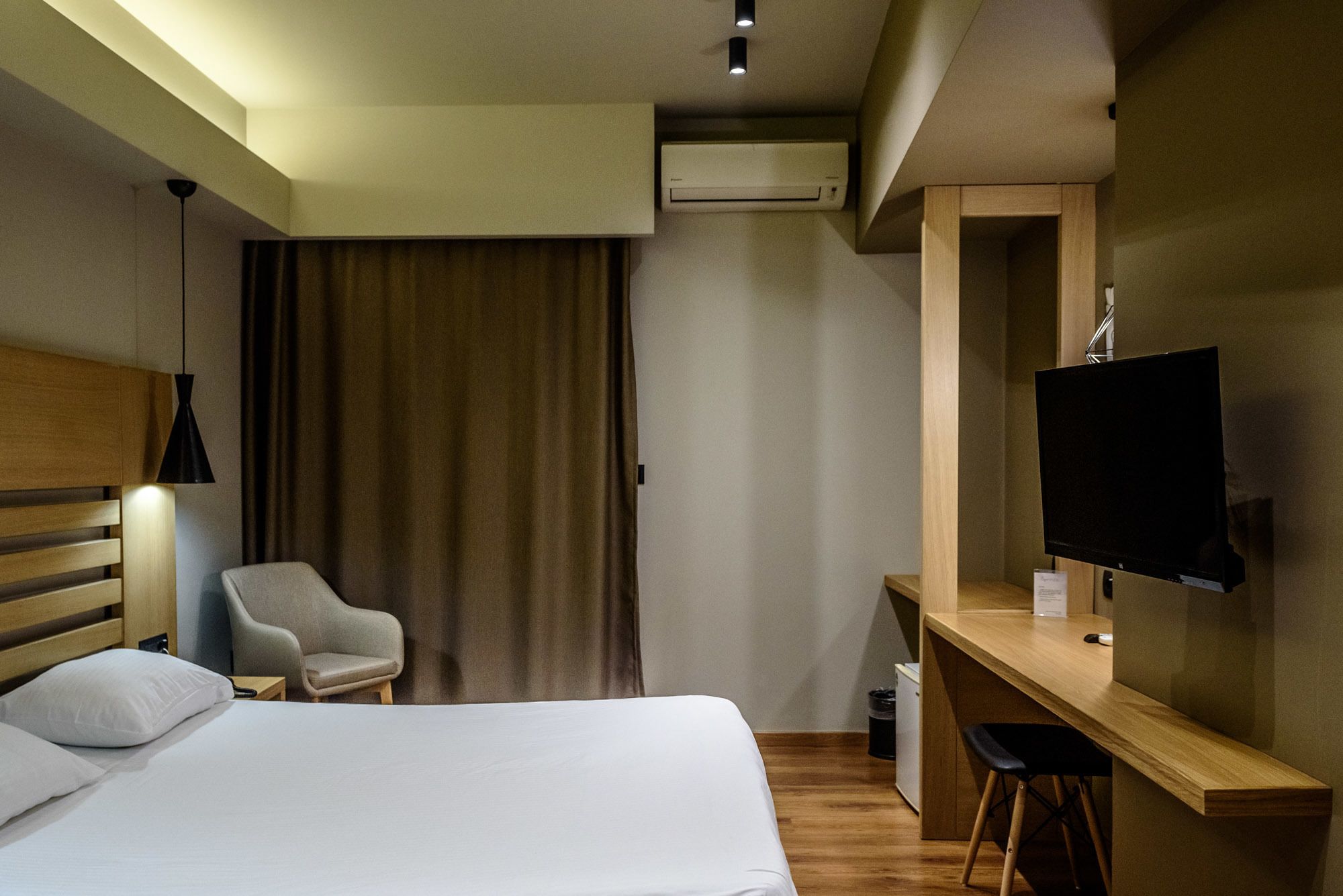 A hotel room with a bed, a desk with a TV, a chair, and an air conditioning unit.