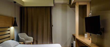 A hotel room with a bed, a desk with a TV, a chair, and an air conditioning unit.