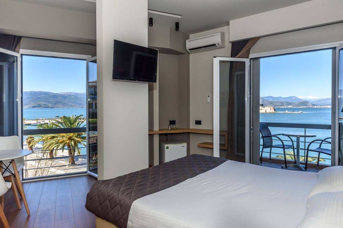 A hotel room with a bed, a TV, a desk, and a balcony with a view of the sea.