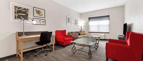 A hotel room with a desk, two red chairs, a glass coffee table, and a TV.
