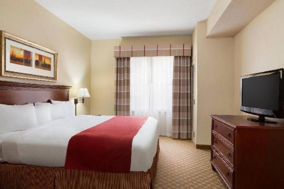 A hotel room with a bed, a television, a dresser, a painting on the wall, and a window with curtains.