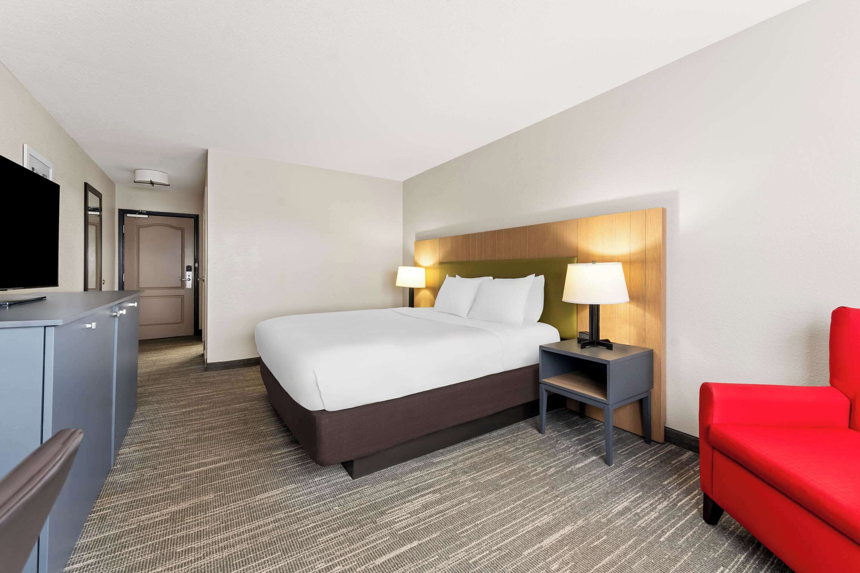 A hotel room with a bed, bedside tables, a TV, a red chair, and a carpeted floor.