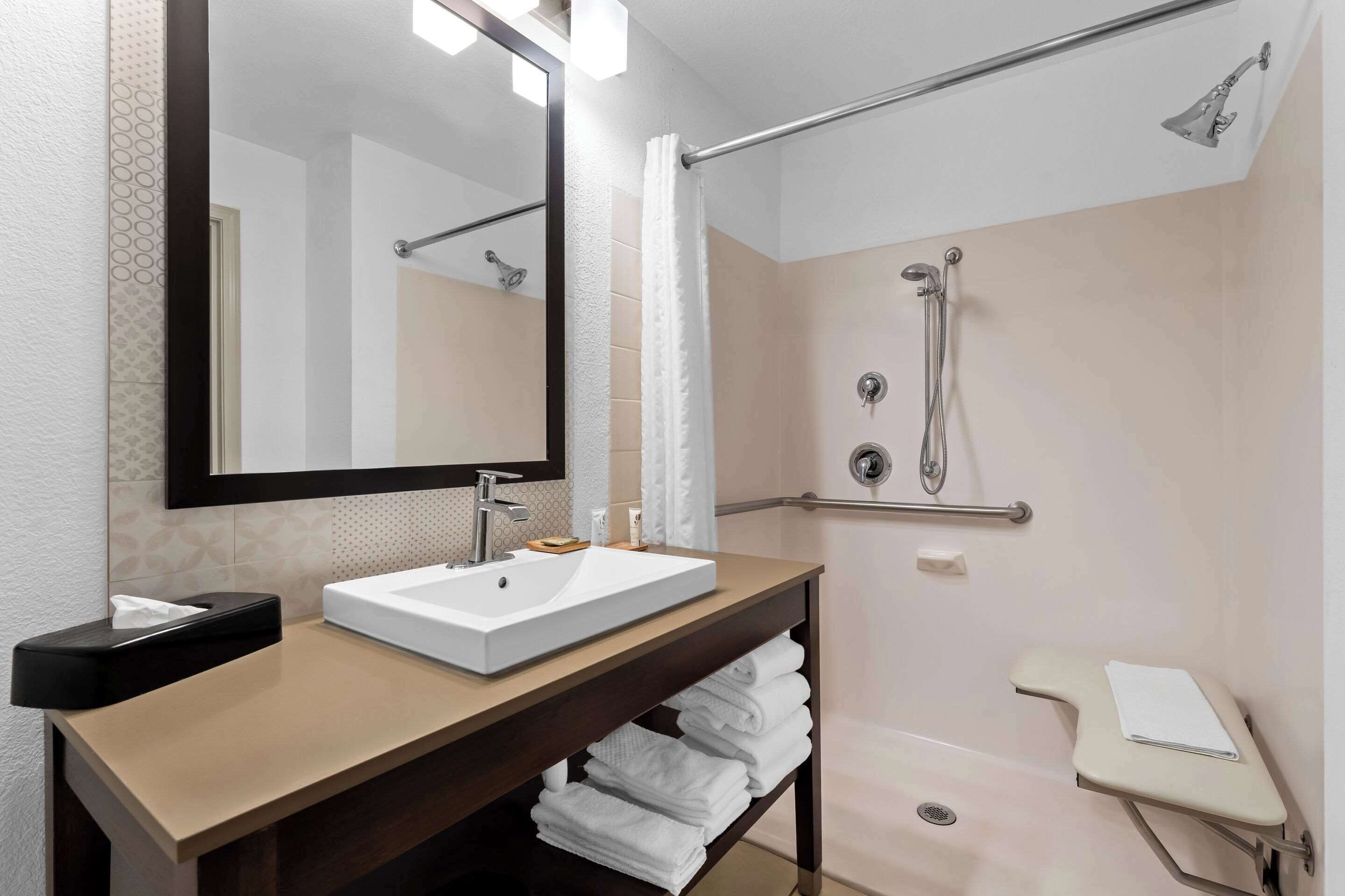 Un baño moderno con un espejo grande, un lavamanos, ducha con barras de apoyo y un perchero para toallas.