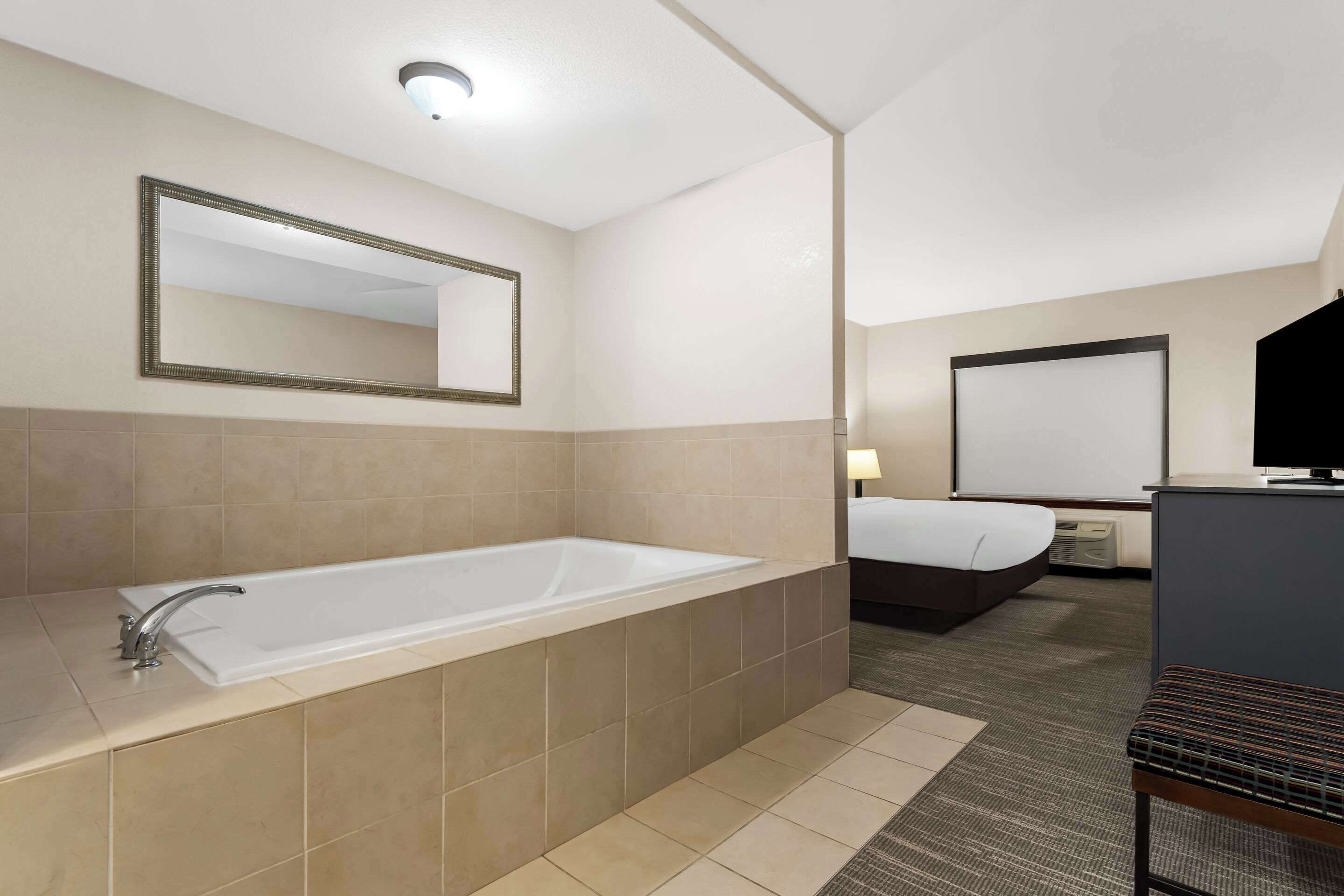 A modern hotel bathroom with a large bathtub, a mirror, and a bed in the adjacent room.