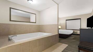 Une salle de bain moderne dans un hôtel, dotée d’une grande baignoire, d’un miroir et d’un lit dans la pièce adjacente.