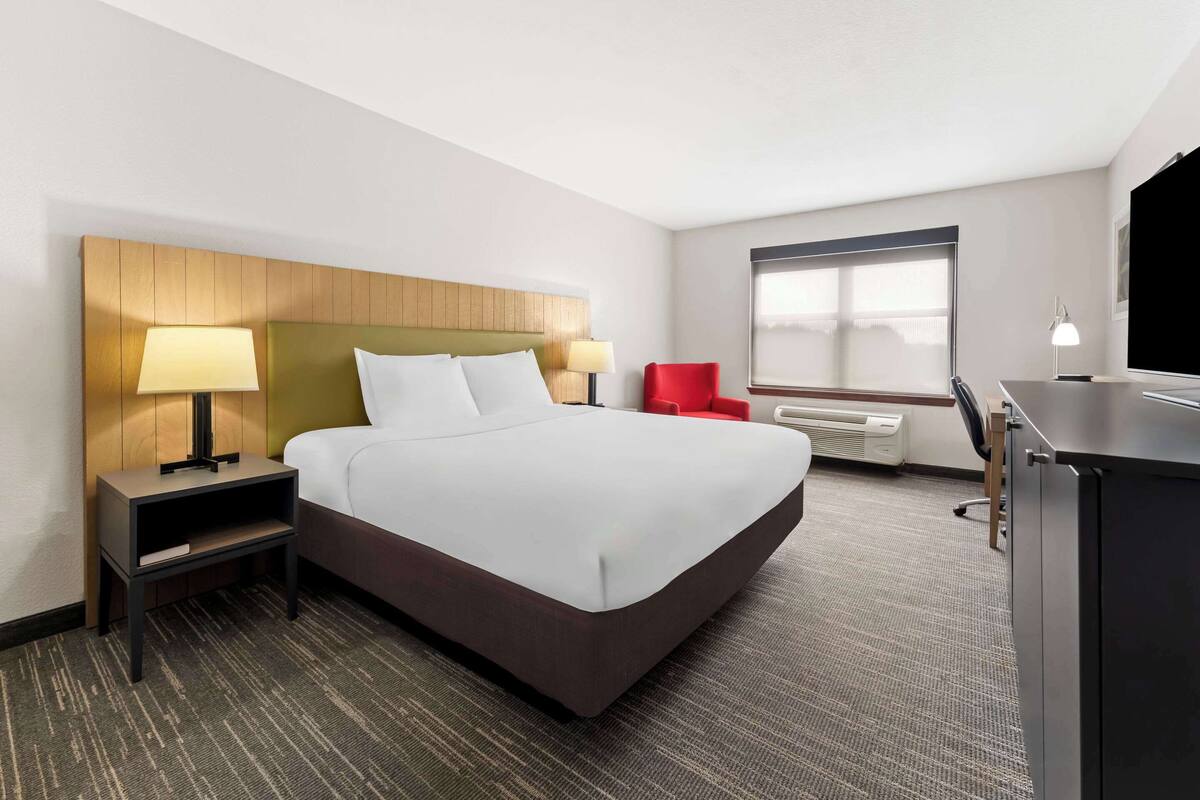 A hotel room with a large bed, two bedside tables with lamps, a red chair, a desk, and a large window.