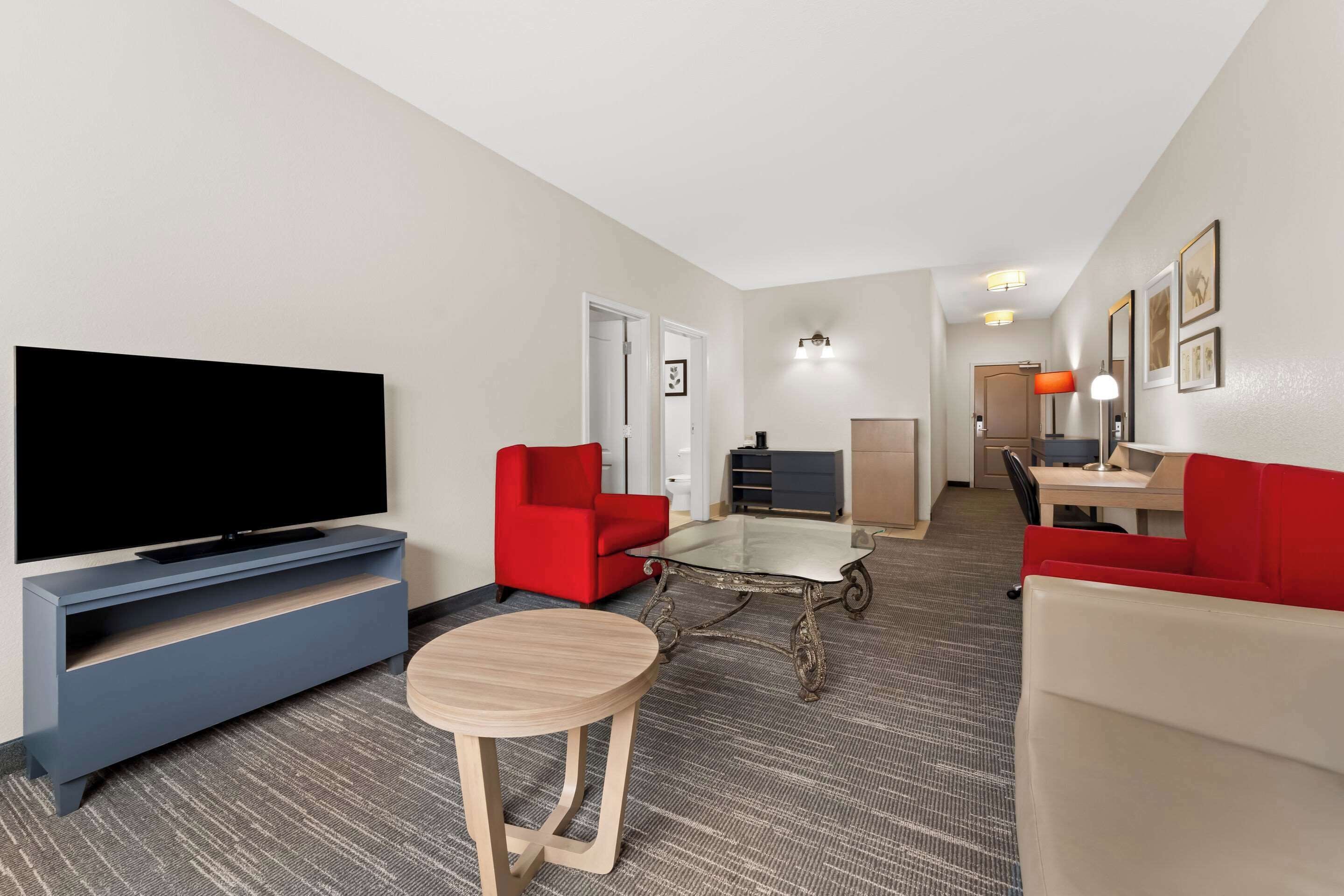 A hotel room with a flat-screen TV, red and white sofas, a small round table, and a glass-top coffee table.