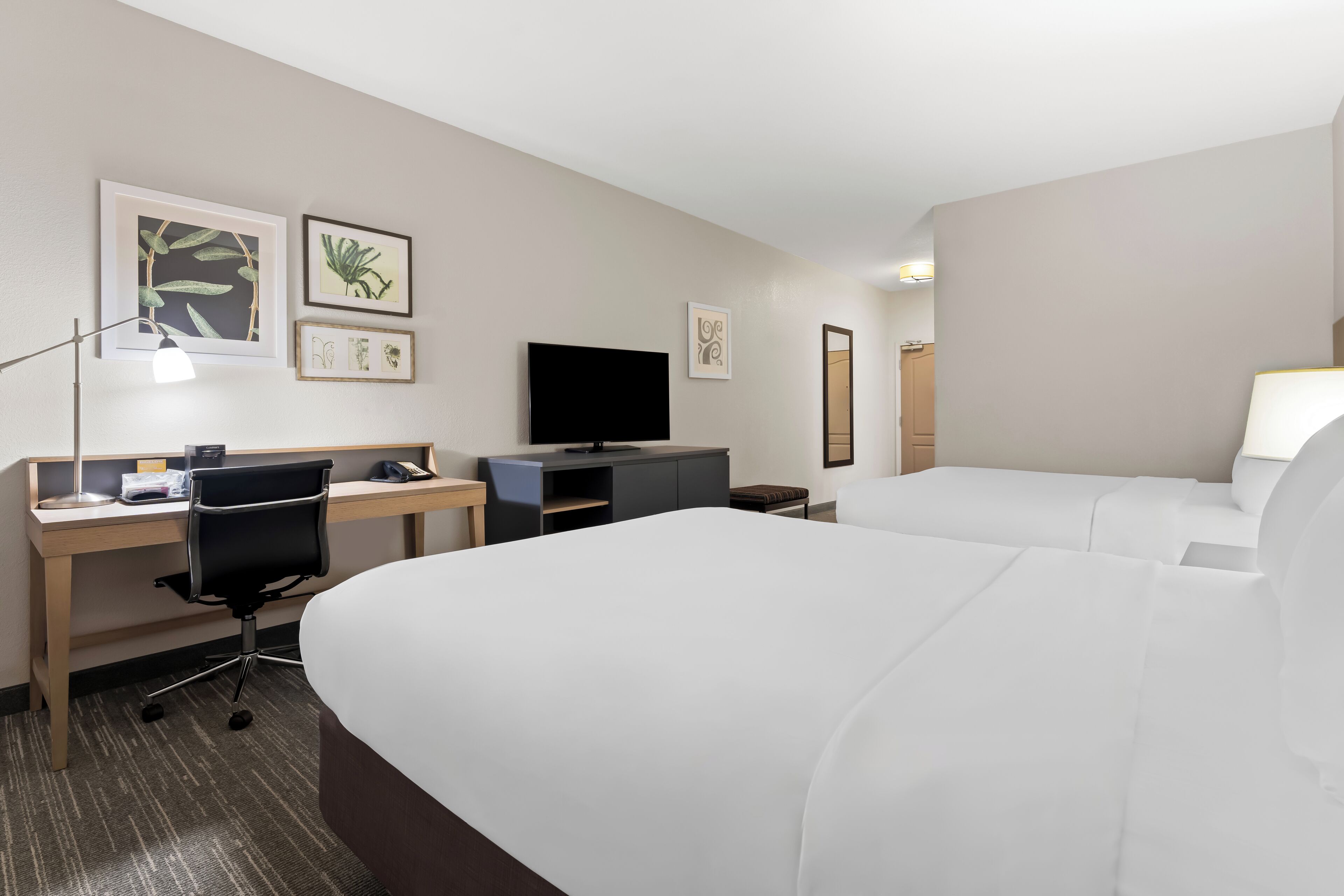 A hotel room with two beds, a desk with a computer, a TV, and framed artwork on the wall.
