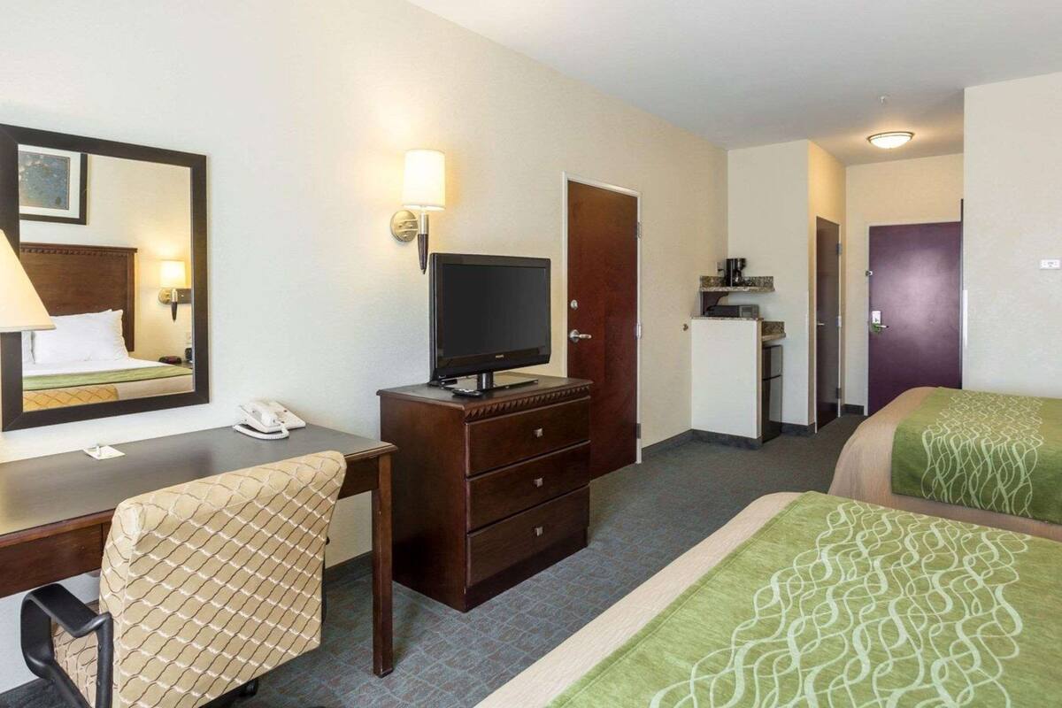 Ett hotellrum med en säng, ett skrivbord med en stol, en tv, en byrå, en spegel och en dörr som leder till ett annat rum.
