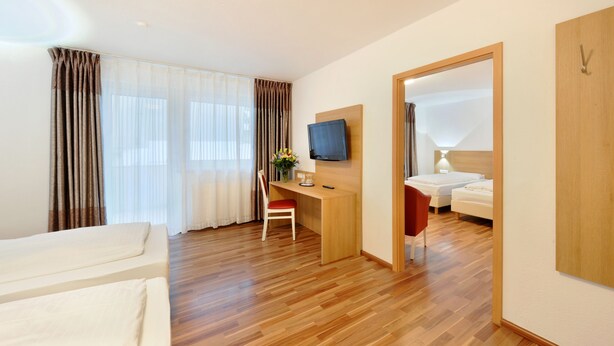 Quarto de hotel com chão de madeira, uma escrivaninha com cadeira, televisão, cama e guarda-roupa.