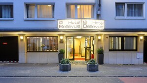 Um hotel chamado "Hotel Bellevue" com uma placa dupla sobre a entrada, o número "98" no prédio e duas plantas em vasos à frente.