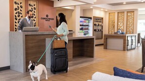 Un espace d’accueil avec un comptoir en bois, un homme et une femme derrière le comptoir, un chien tenu en laisse et un chariot à bagages.