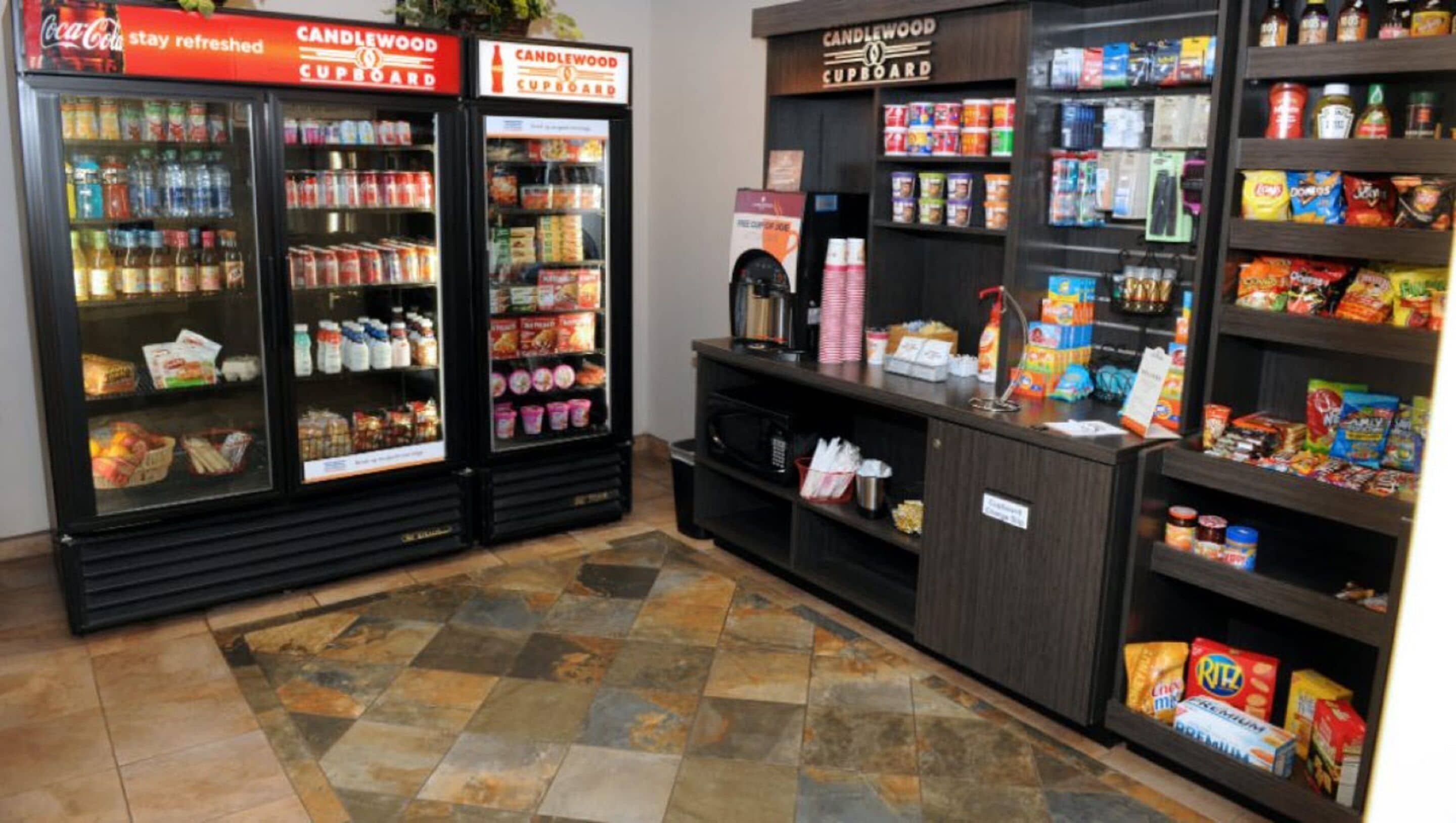 A self-service beverage and snack area with refrigerated drink dispensers, a coffee machine, and shelves stocked with various snacks and beverages.
