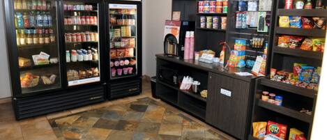 Uma área de autoatendimento com bebidas e lanches, incluindo máquinas de refrigerantes, cafeteira e prateleiras repletas de diversas opções de lanches e bebidas.
