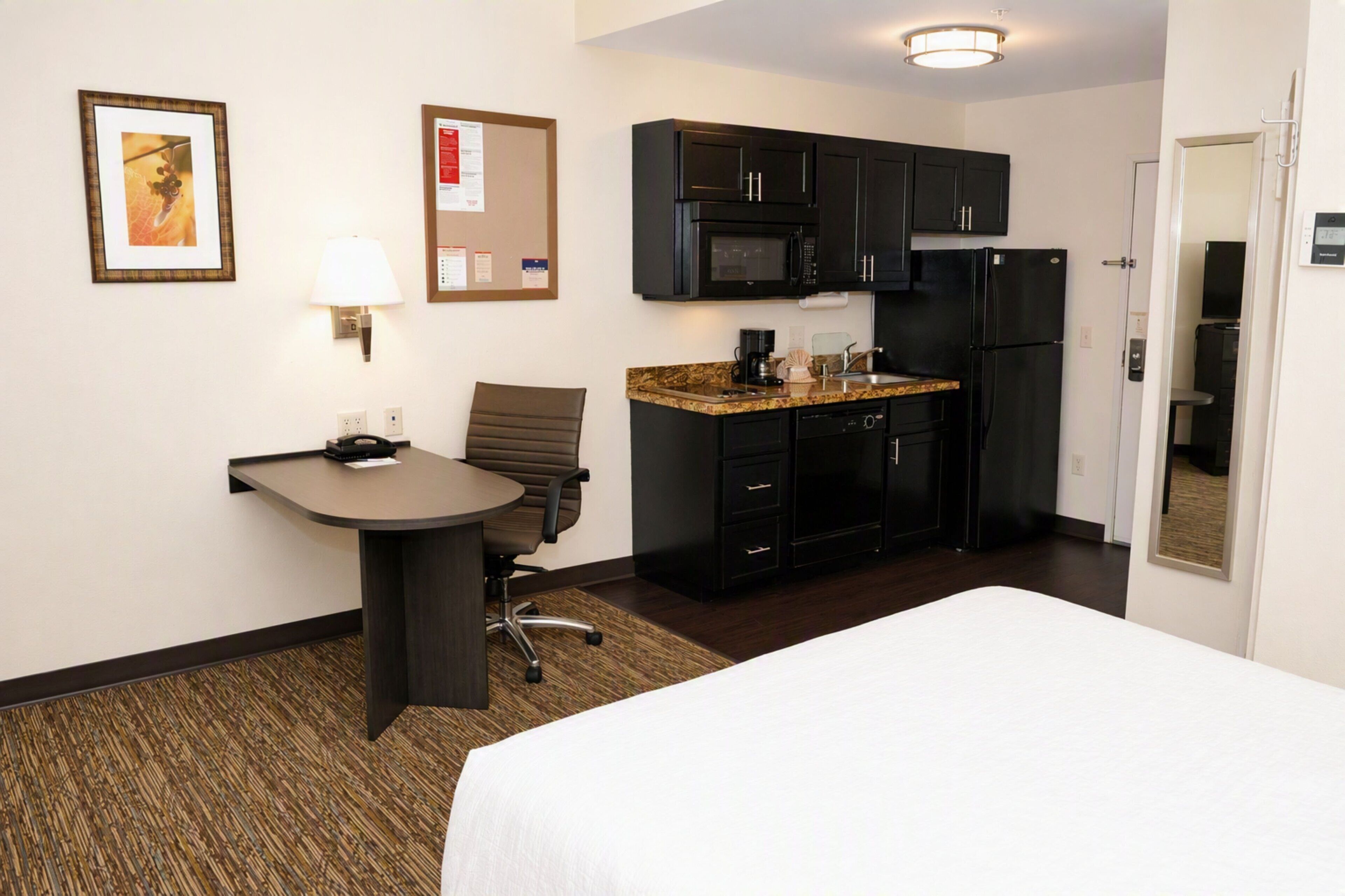 A hotel room with a bed, a desk, a chair, a microwave, a refrigerator, and a small kitchenette.