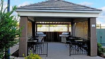 A covered outdoor patio with two barbecue grills, a concrete floor, and a metal fence.
